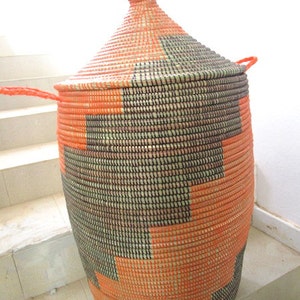 May include: A large, woven storage basket with a lid. The basket is made of natural fibers and is decorated with a geometric pattern in orange and black.