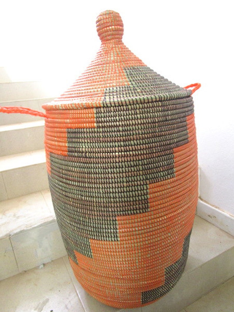 May include: A large, woven storage basket with a lid. The basket is made of natural fibers and is decorated with a geometric pattern in orange and black.