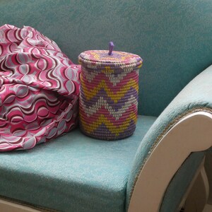 May include: A pink and white patterned fabric sits on a teal blue sofa with a white wooden armrest. A woven basket with a purple lid and pink, yellow, and white stripes sits on the sofa.