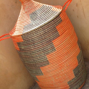 May include: A large, orange and brown woven basket with a lid and handles. The basket is made of natural materials and has a traditional design.