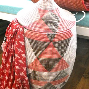 May include: A large, woven storage basket with a lid. The basket is made of natural materials and has a geometric pattern of black, white, and pink triangles. The basket is sitting on a wooden floor.