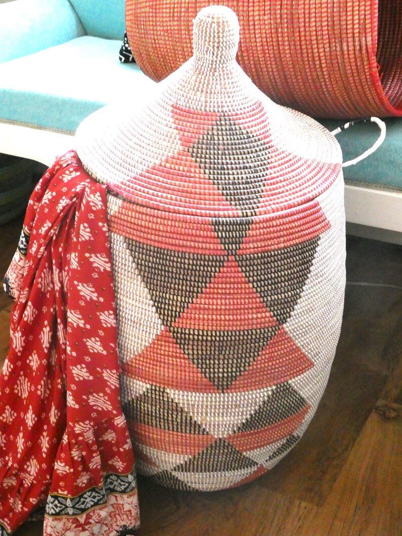 May include: A large, woven storage basket with a lid. The basket is made of natural materials and has a geometric pattern of black, white, and pink triangles. The basket is sitting on a wooden floor.