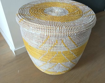 Puede incluir: Una cesta de almacenamiento grande, redonda y tejida con tapa. La cesta está hecha de materiales naturales y está pintada con un patrón geométrico amarillo y blanco.