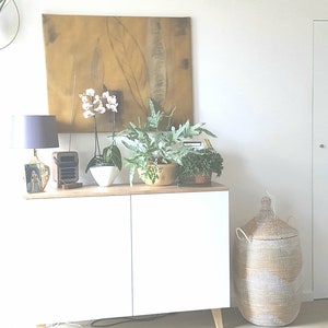 May include: A white cabinet with two doors and wooden legs sits on a light wood floor. The cabinet is decorated with a lamp, a radio, and three potted plants. A large woven basket sits on the floor to the right of the cabinet.