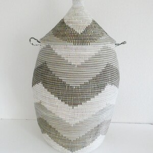 May include: An oval-shaped woven storage basket with a lid. The basket has a geometric pattern of white, grey, and black stripes. The lid is conical with a small handle. The basket is made of natural materials and is suitable for storage.