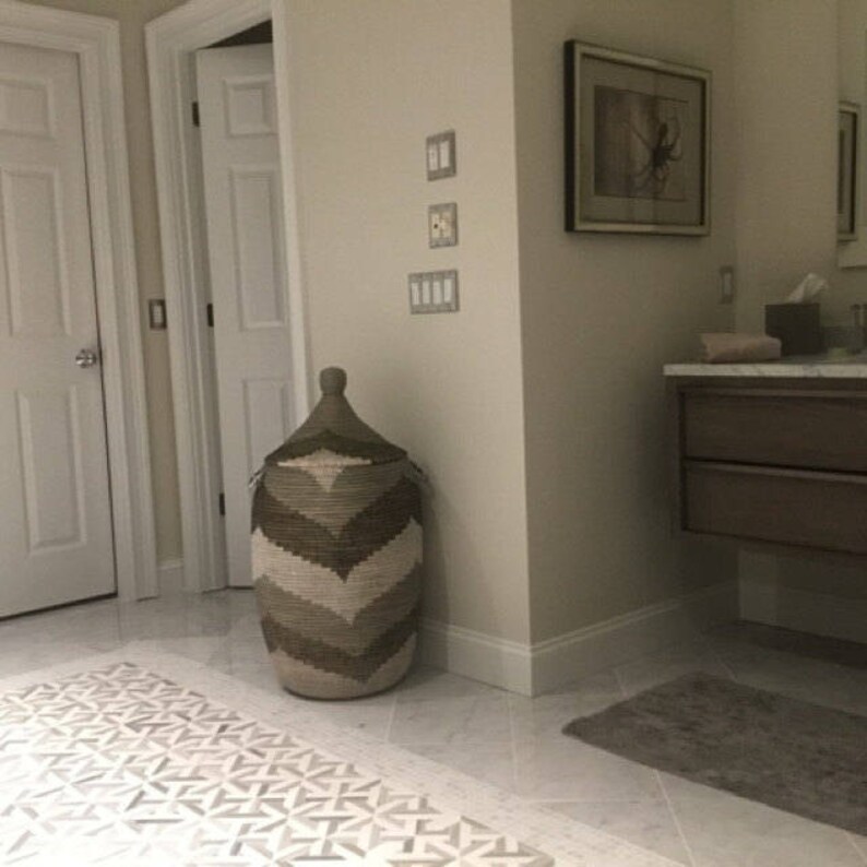 May include: A tall, woven storage basket with a conical lid. The basket features a chevron pattern in shades of brown and beige. It is placed in a bathroom setting, next to a wall and a patterned rug.