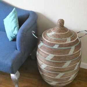 May include: A large, woven storage basket with a lid and handles. The basket is made of natural brown and white fibers and has a geometric pattern. The basket is sitting on a wooden floor next to a blue couch.