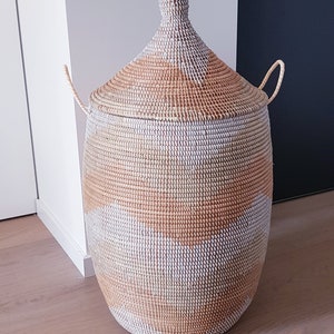 May include: A large, woven storage basket with a lid. The basket is made of natural materials and has a white and brown geometric pattern. The basket has two handles on the sides.