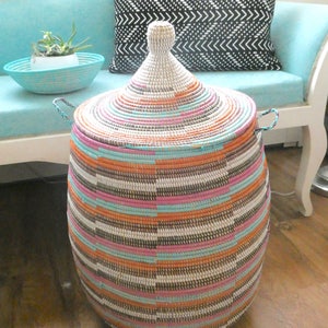 May include: A large, round, woven storage basket with a lid. The basket is made of natural materials and has a colorful striped pattern in shades of pink, orange, blue, and white. The lid is domed and has a similar striped pattern.