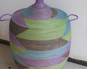 May include: A large, woven storage basket with a lid. The basket is made of natural fibers and is painted in shades of purple, green, and blue. The basket has a unique geometric pattern.