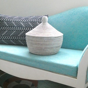 May include: A white wicker basket with a lid sits on a turquoise blue couch with a black and white patterned pillow. The couch has white painted legs.