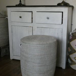 Op de afbeelding: Een witte rieten opbergmand met een deksel. De mand is rond en heeft een geweven patroon. Hij staat voor een witte kast met twee lades.
