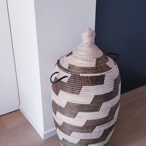 May include: A woven storage basket with a lid. The basket is oval-shaped and features a geometric pattern in shades of brown and cream. It has two black handles and a decorative top.