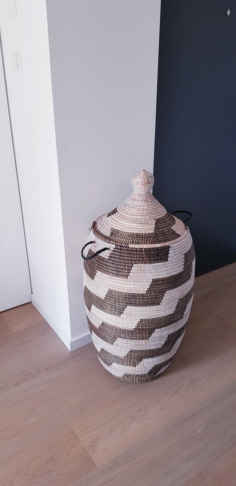 May include: A woven storage basket with a lid. The basket is oval-shaped and features a geometric pattern in shades of brown and cream. It has two black handles and a decorative top.