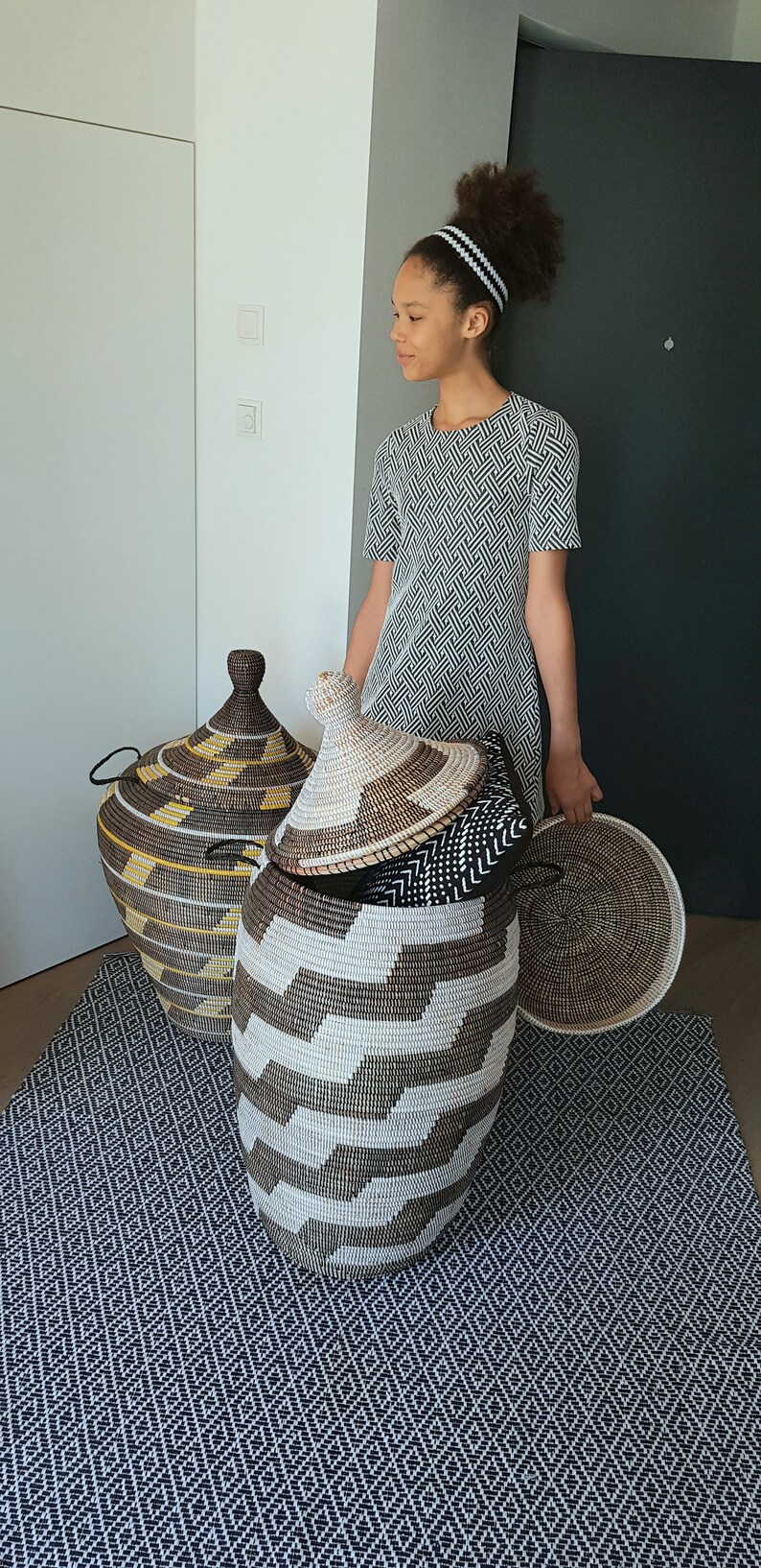 May include: Woven storage baskets in various sizes and patterns. The largest basket is open, revealing smaller baskets and a lid. The baskets are brown, white, and yellow. The scene is set on a patterned rug.