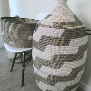 May include: Two woven storage baskets. The larger basket has a lid and is cylindrical with a geometric pattern in white and dark brown. The smaller basket is oval and sits on a white chair. Both baskets are made of natural fibers.