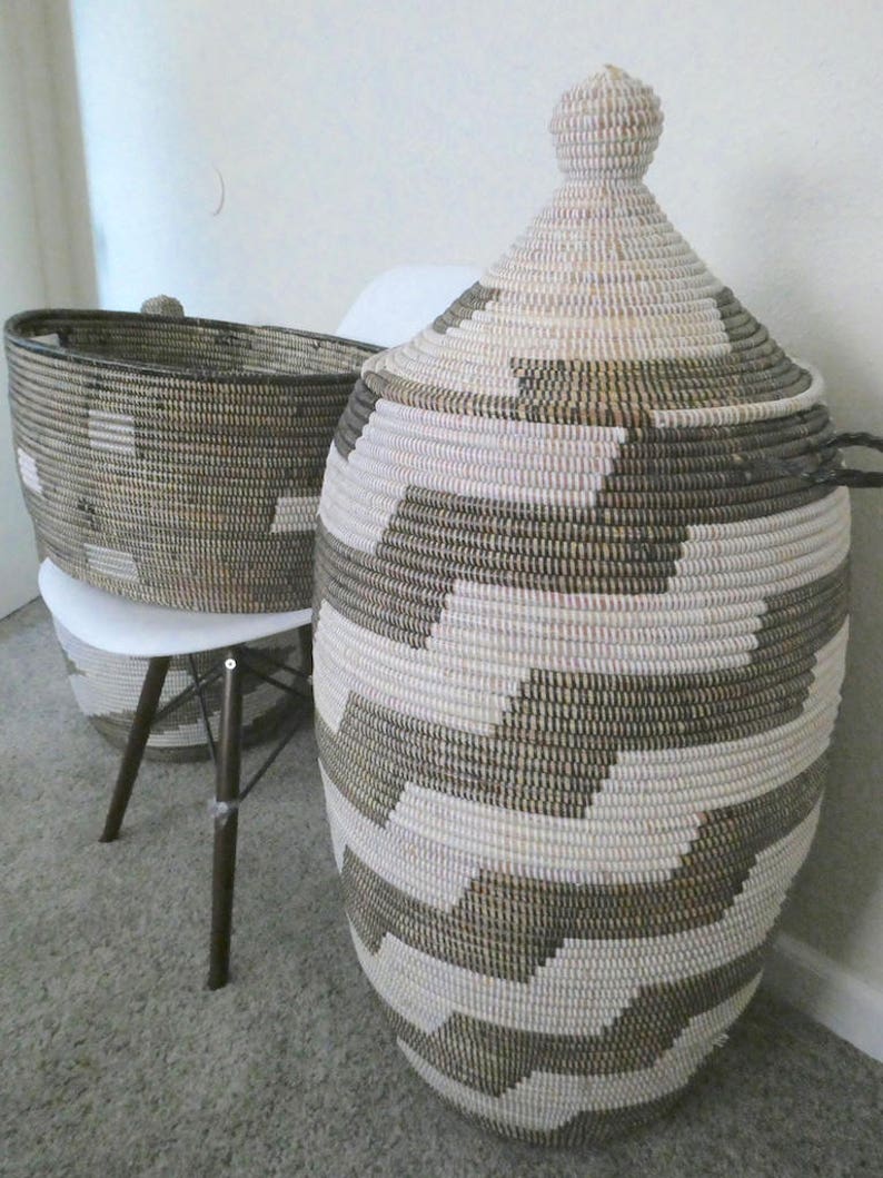 May include: Two woven storage baskets. The larger basket has a lid and is cylindrical with a geometric pattern in white and dark brown. The smaller basket is oval and sits on a white chair. Both baskets are made of natural fibers.