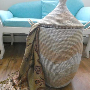 May include: A large white and brown woven storage basket with a lid and handles. The basket is sitting on a wooden floor next to a light blue couch.