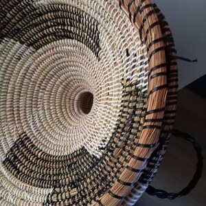 May include: Woven basket with a circular design. The basket features a spiral pattern in natural, black, and white tones. The rim and handle are black. The basket is made of natural fibers.