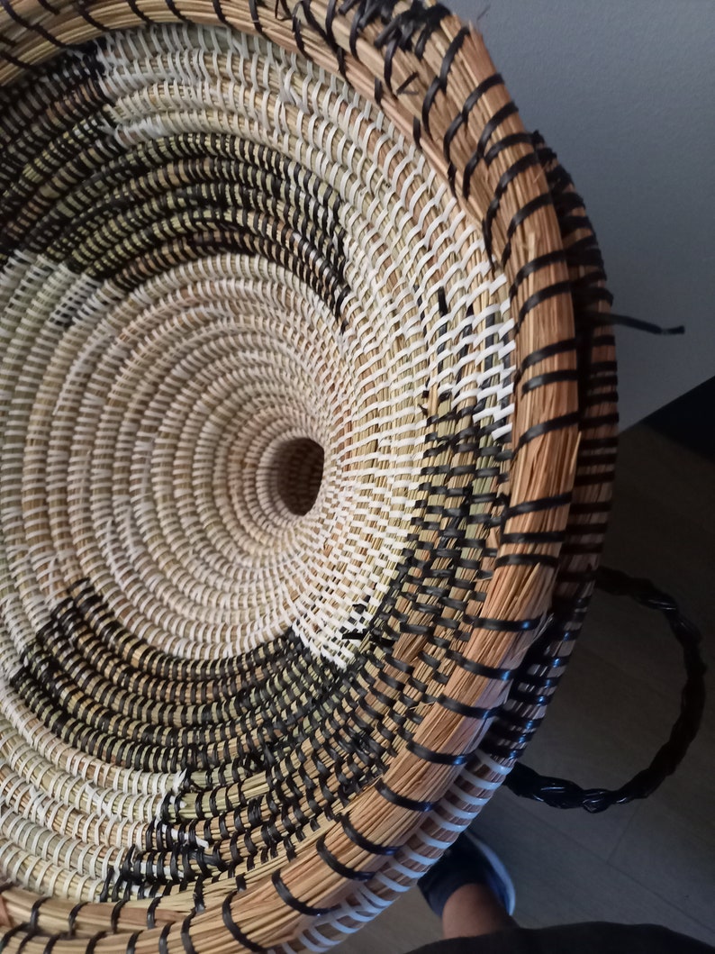 May include: Woven basket with a circular design. The basket features a spiral pattern in natural, black, and white tones. The rim and handle are black. The basket is made of natural fibers.