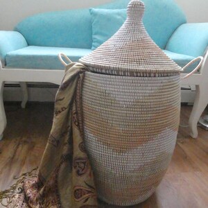 May include: A white and brown woven storage basket with a lid and handles. The basket is sitting on a wooden floor next to a light blue couch.