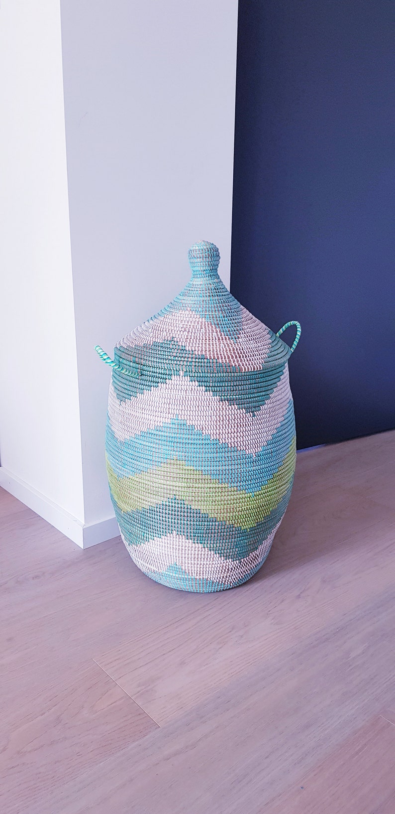 May include: A large, woven storage basket with a lid. The basket is made of natural materials and has a blue, green, and white geometric pattern.
