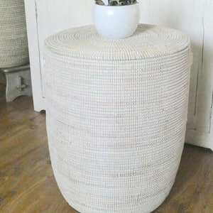 Op de afbeelding: Een witte geweven opbergmand met een deksel, staand op een houten vloer. Een kleine witte pot met een vetplant staat op de mand.