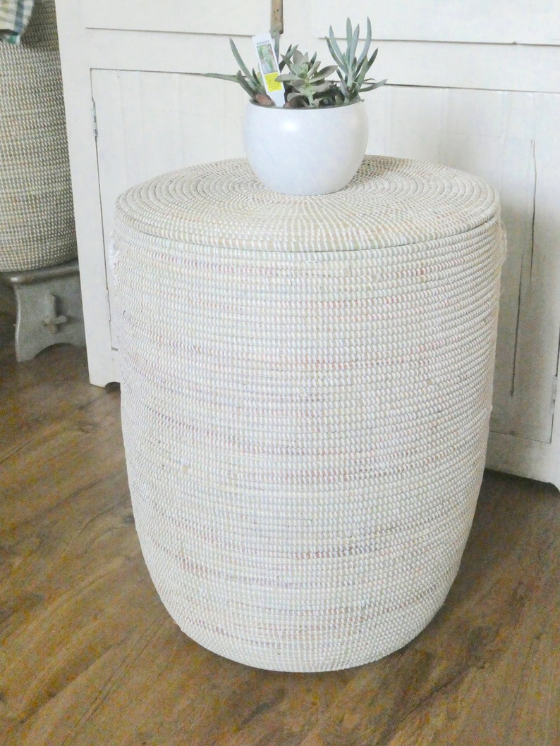 Op de afbeelding: Een witte geweven opbergmand met een deksel, staand op een houten vloer. Een kleine witte pot met een vetplant staat op de mand.