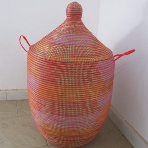 May include: A large, woven storage basket with a lid. The basket is made of natural fibres and is painted in shades of pink, orange, and gold. The lid has a rounded top and two red handles.