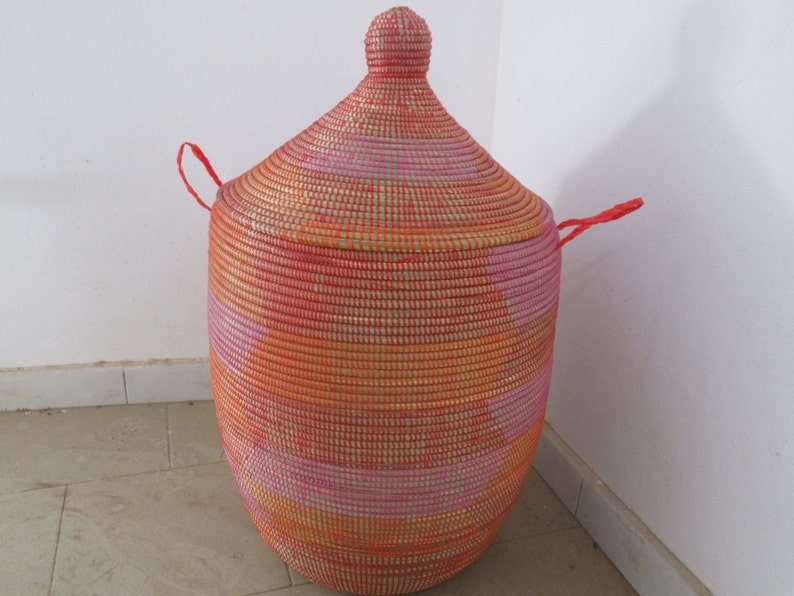May include: A large, woven storage basket with a lid. The basket is made of natural fibres and is painted in shades of pink, orange, and gold. The lid has a rounded top and two red handles.
