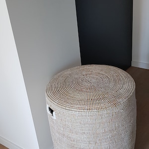 Op de afbeelding: Een ronde, geweven opbergmand van wit en bruin met een deksel. De mand heeft een handvat aan de zijkant.