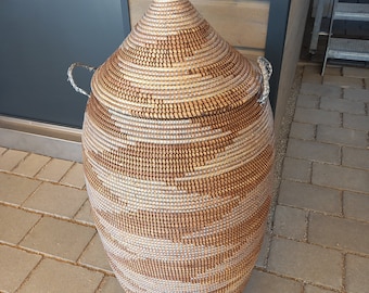 May include: A large, woven storage basket with a lid. The basket is made of natural materials and has a brown and white pattern. The basket is sitting on a brick patio.
