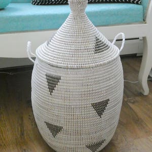 May include: A white woven storage basket with a lid. The basket has a rounded shape with a conical lid and two small handles. Black triangle patterns are spaced around the basket. The basket is sitting on a wooden floor.