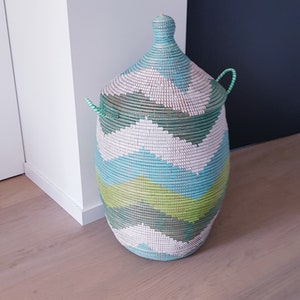 May include: A large, woven storage basket with a lid. The basket is green, blue, and white with a chevron pattern. The basket is sitting on a wooden floor.