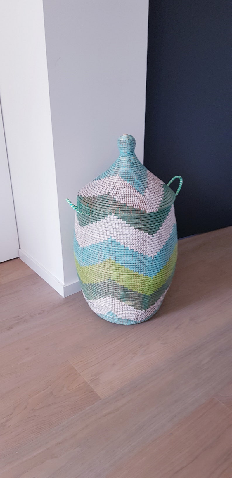 May include: A large, woven storage basket with a lid. The basket is green, blue, and white with a chevron pattern. The basket is sitting on a wooden floor.