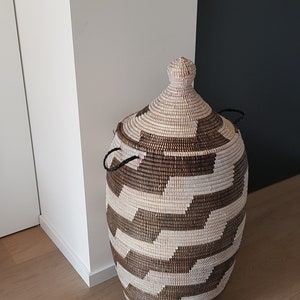 May include: A woven storage basket with a lid. The basket is cylindrical with a rounded top and is made of natural and brown fibers. The lid has a knob and two black handles. The basket is placed on a wooden floor.