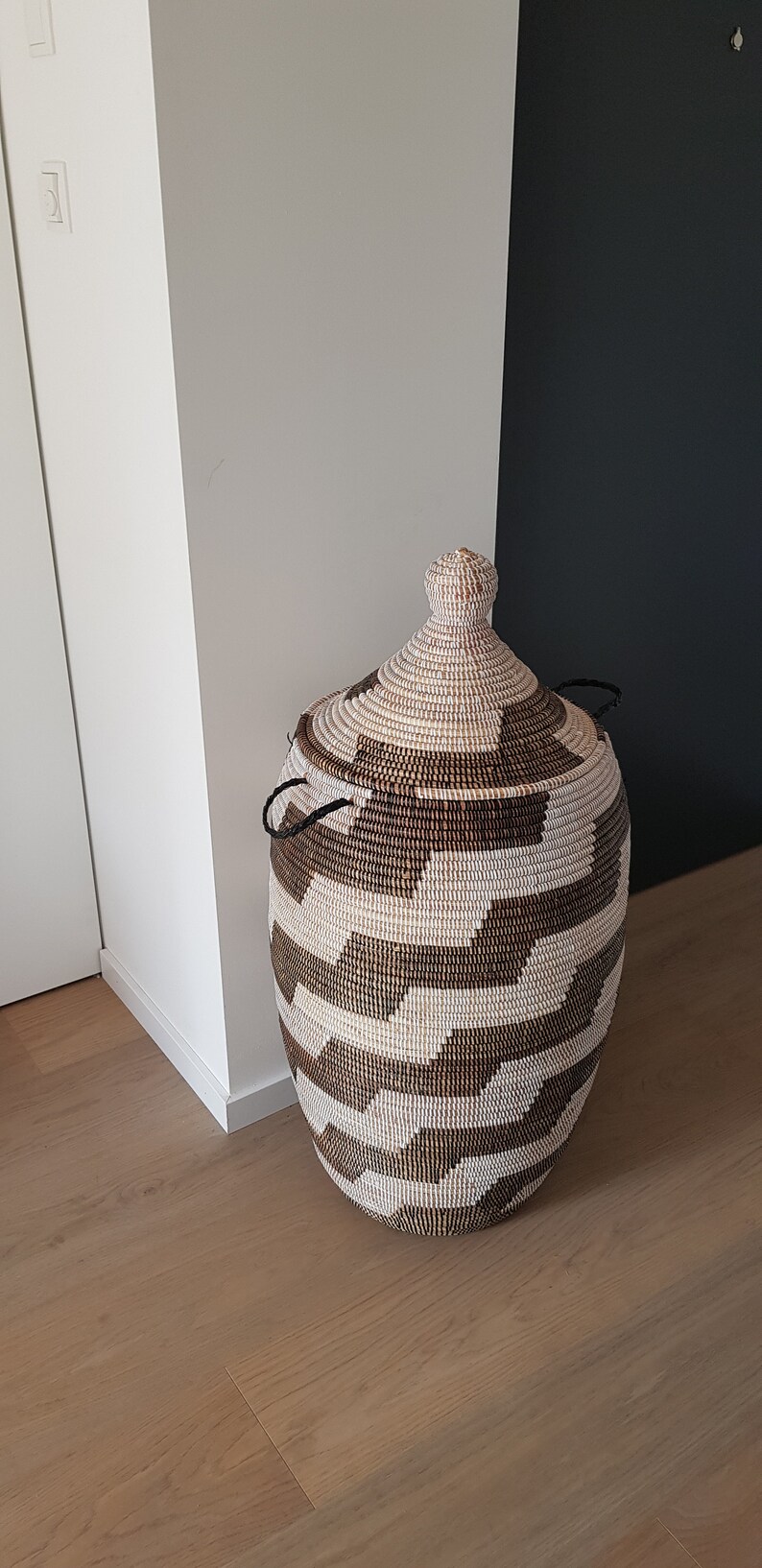 May include: A woven storage basket with a lid. The basket is cylindrical with a rounded top and is made of natural and brown fibers. The lid has a knob and two black handles. The basket is placed on a wooden floor.