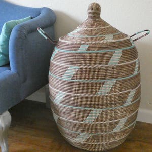 May include: A large, woven storage basket with a lid and handles. The basket is brown with white and light blue stripes. It is sitting on a wooden floor next to a blue couch.