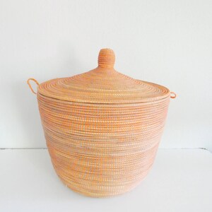 May include: A large, round, woven storage basket with a lid. The basket is made of natural materials and has a light brown color with hints of orange. The lid has a small knob on top.