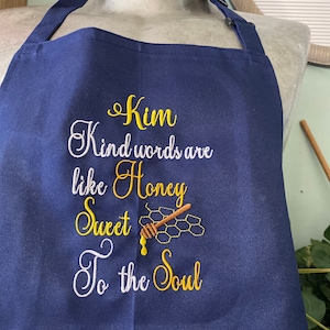 May include: A navy blue apron with the text "Kim Kind words are like Honey Sweet To the Soul" embroidered in yellow thread. A small honey dipper and honeycomb are also embroidered on the apron.