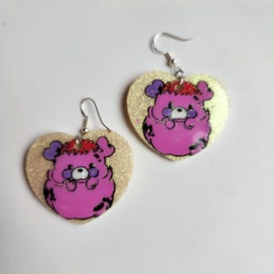 May include: A pair of heart-shaped earrings with a pink cartoon character on each. The earrings are made of gold glitter resin and have silver hooks.