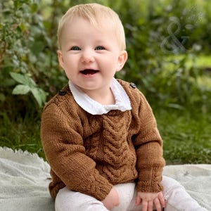 Puede incluir: Un niño pequeño viste un suéter de punto trenzado marrón de manga larga con una camisa de cuello blanco debajo. El suéter presenta un patrón de trenzas vertical en la parte delantera y botones oscuros en los hombros. El niño lleva pantalones claros.