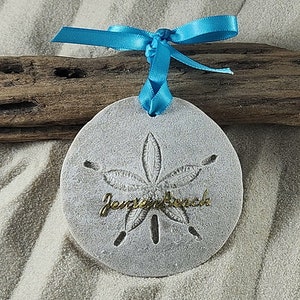 May include: A white sand dollar ornament with a gold inscription that reads "Jensen Beach". The ornament is tied with a blue ribbon.