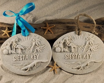 May include: Two white sand-textured oval ornaments with a raised image of palm trees, a sun, and flamingos. The text "SIESTA KEY" is embossed on each ornament. One ornament has a blue ribbon attached, and the other has a brown twine string.