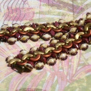 Puede incluir: Una pulsera de cuentas con un diseño geométrico. La pulsera incorpora cuentas de color burdeos, dorado y crema. El diseño incluye formas de diamante, media luna y redondas, con un cierre de color bronce.