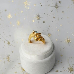 May include: A gold ring with a blue gemstone and a detailed fox design. The ring is displayed in a white ring box.