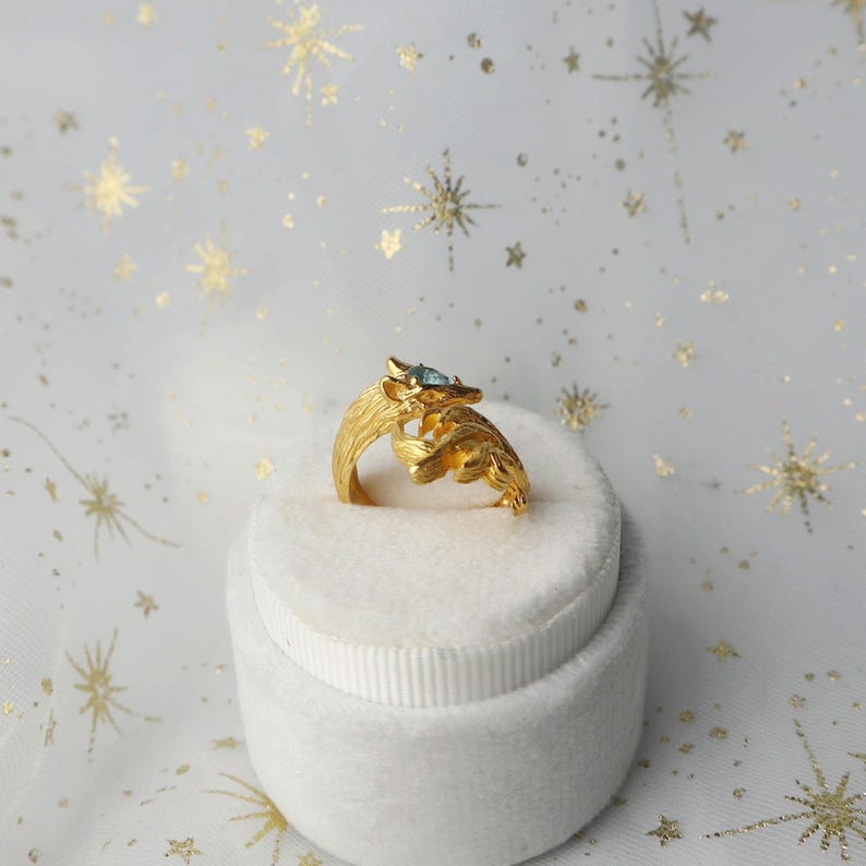 May include: A gold ring with a blue gemstone and a detailed fox design. The ring is displayed in a white ring box.