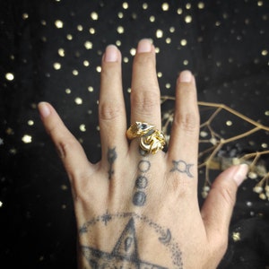 May include: A gold ring with a detailed design of two foxes facing each other. The ring is worn on the middle finger of a hand with several tattoos.