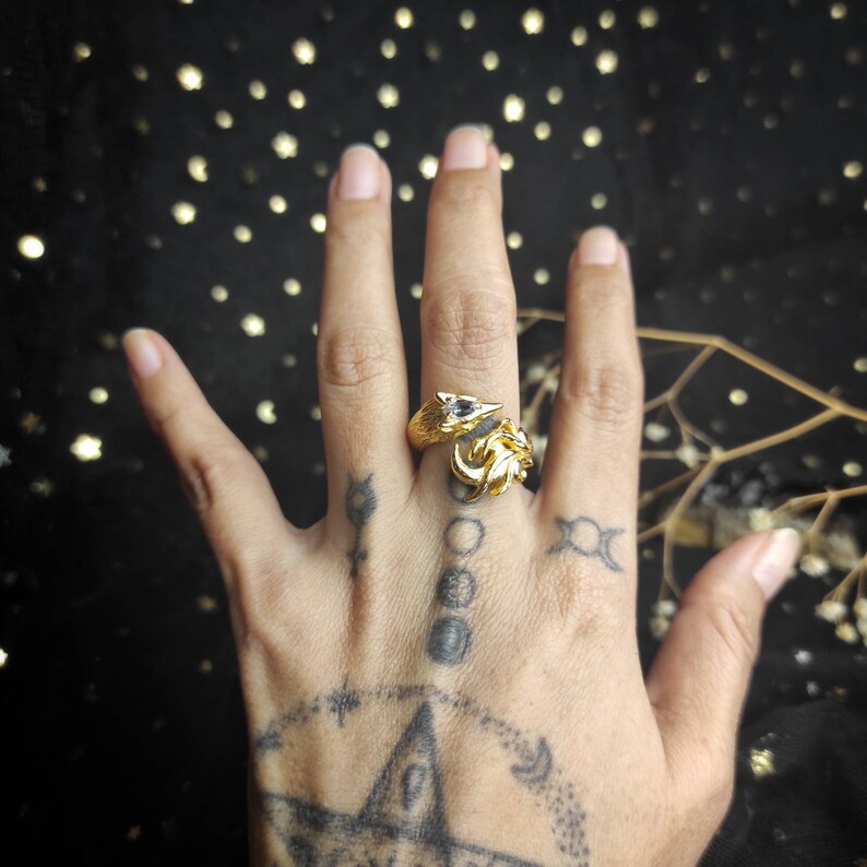 May include: A gold ring with a detailed design of two foxes facing each other. The ring is worn on the middle finger of a hand with several tattoos.
