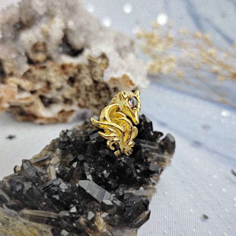 May include: A gold-colored fox figurine with a pink gemstone eye, sitting on a bed of black crystals.
